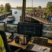 Control room overlooking a canal with a crowded bridge race visible outside; multiple monitors show canal views and control data; high‑visibility vest reads BRUGWACHTER in the foreground.