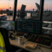 Bridge operator station at sunset with multiple screens displaying canal locks and controls; high‑visibility vest labeled BRUGWACHTER draped over chair.
